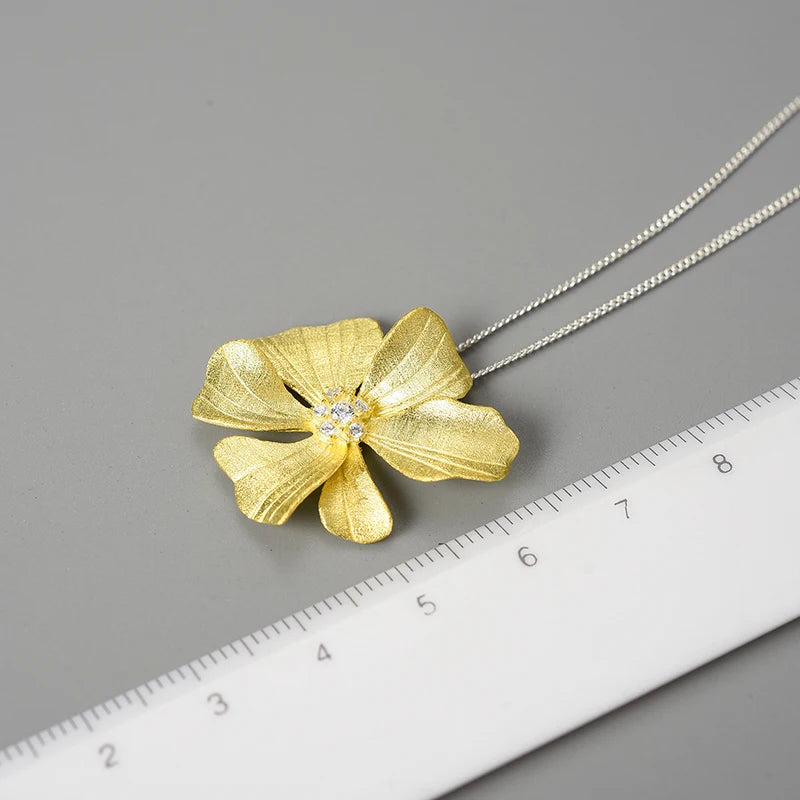 A textured yellow gold flower pendant necklace with a small clear crystal center, shown on a silver chain next to a white ruler for scale on a gray background.